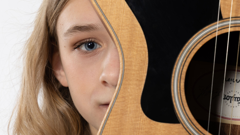 Close-up van een kind met blond haar dat achter een akoestische gitaar vandaan kijkt, met één blauw oog zichtbaar