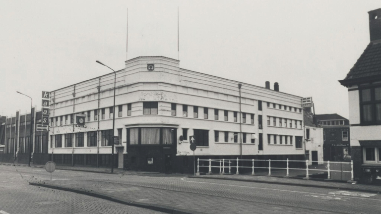 Oud zwart-wit-foto van een hoekig gebouw met een bord "kaas" en een klok aan de gevel; straten zijn geplaveid.