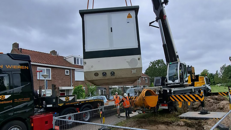 Kraanwagen hijst een grote prefab-unit boven een bouwplaats, met twee bouwvakkers in oranje hesjes erbij