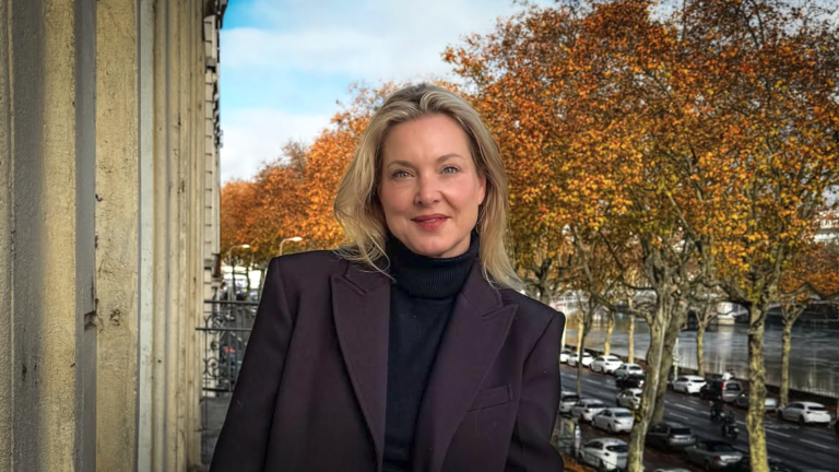 Vrouw met blond haar poseert buiten voor bomen met herfstbladeren en een straat op de achtergrond.