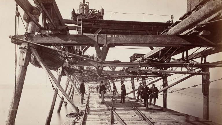 Houten steigerconstructie boven water met mannen en spoorrails, waarschijnlijk tijdens bouwactiviteiten eind 19e eeuw.
