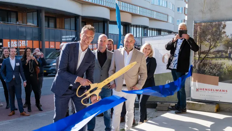 Mensen in nette kleding voeren een lintjesknipceremonie uit met een grote gouden schaar voor een nieuw gebouw, terwijl toeschouwers foto's maken.