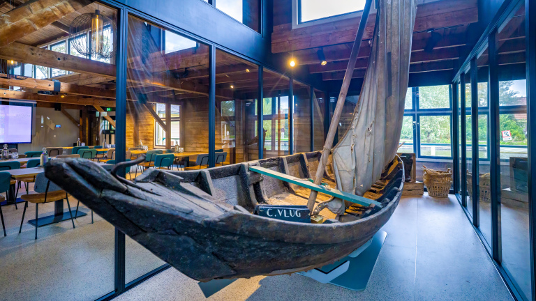 Oude houten zeilboot als decorstuk in een modern gebouw met veel glas, houten balken en tafels met stoelen