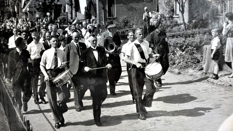 Optocht van een muziekgezelschap met trommelaars en blazers op een zonnige dag, omringd door mensen langs de straat.