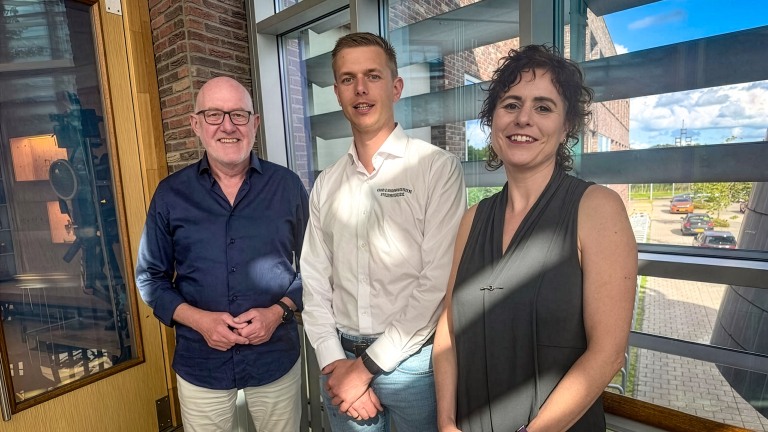 Drie mensen poseren en glimlachen bij een groot raam in een kantoorgebouw, met parkeerplaats en lucht op de achtergrond
