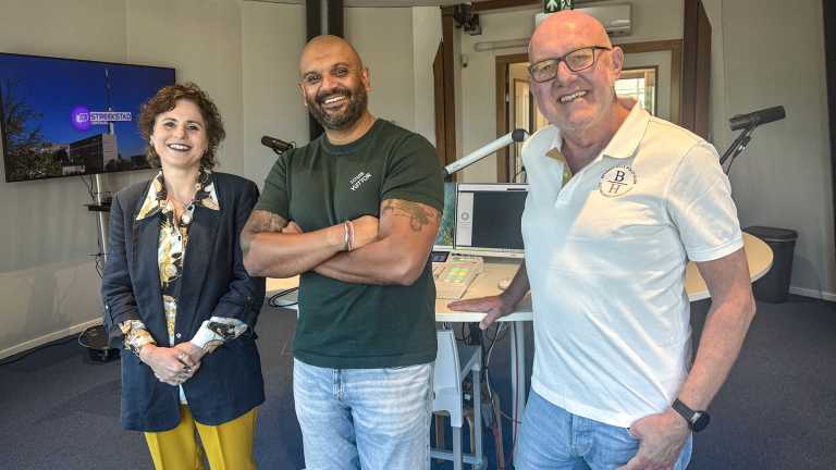 Drie lachende mensen in een radiostudio, met microfoons en een scherm met het logo StadsStreekstad Centraal
