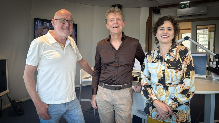 Drie mensen poseren en glimlachen in een radiostudio, met microfoonarm en computers op de achtergrond