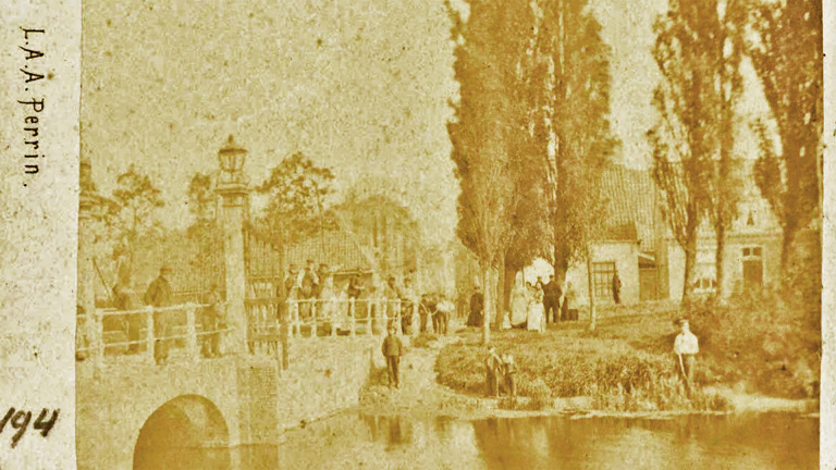 Mensen wandelen over een brug naast een waterpartij, met bomen en gebouwen op de achtergrond in een historische setting.