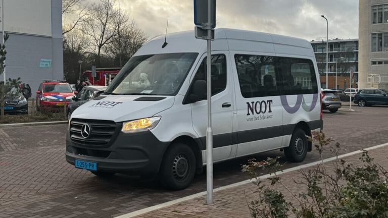 Witte Mercedes-bus met logo 'NOOT' geparkeerd op een straat met bomen en een gebouw op de achtergrond.