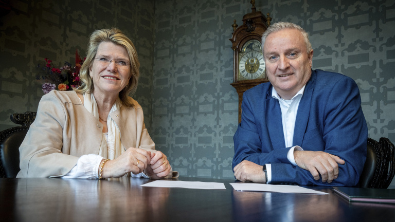 Een man en vrouw zitten aan een tafel met een klok op de achtergrond, beiden glimlachend in een formele setting.