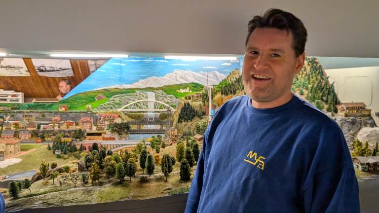 Man in blauwe trui poseert voor een gedetailleerde modelspoorbaan met bergen, huizen en brug.