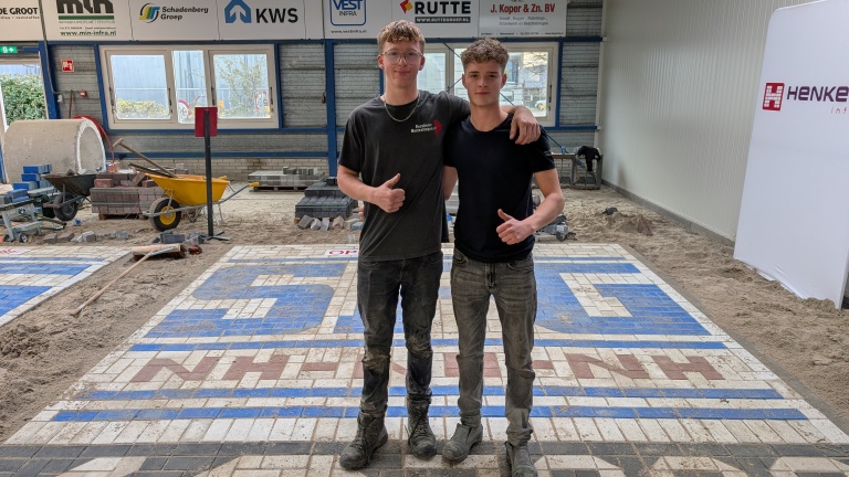 Twee jonge mannen staan zij aan zij met hun armen over elkaars schouders in een ruimte met bestrating in constructie. Ze geven allebei een duim omhoog en dragen werkkleding bedekt met stof. Op de achtergrond is bouwmateriaal zichtbaar, zoals een kruiwagen en stapels stenen, onder borden met bedrijfslogo's.