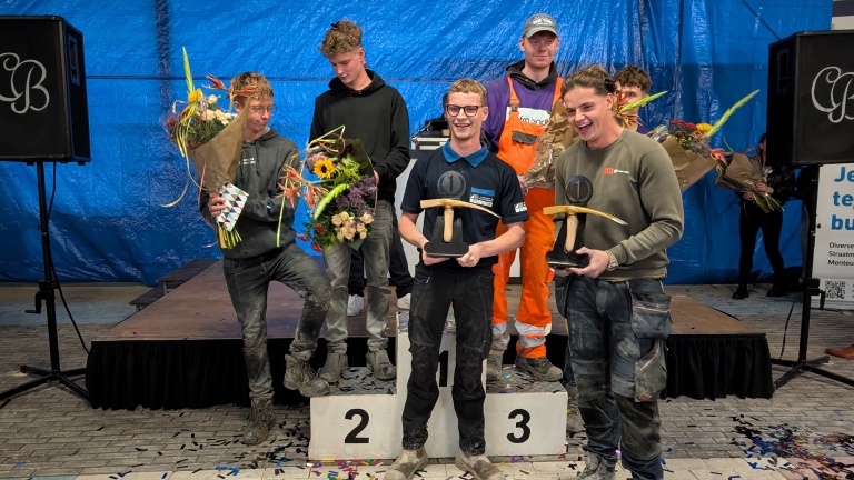 Een groep mannen poseert op een podium; twee van hen houden een prijs vast en anderen hebben bloemen in hun hand.