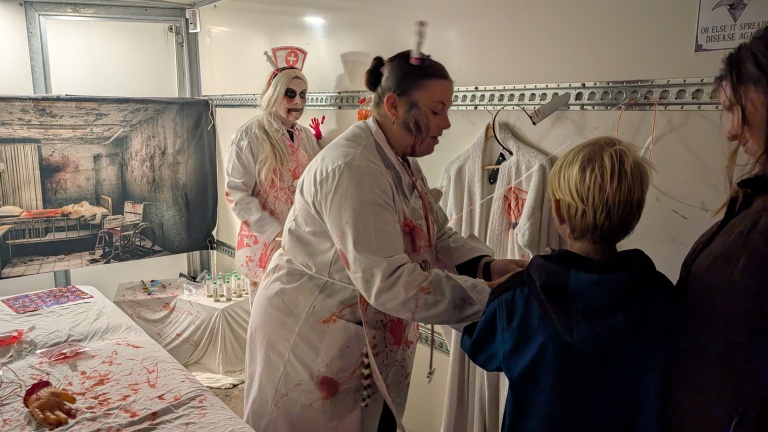 Twee vrouwen verkleed als bloederige verpleegsters in een enge kamer decor met een kind. Eén verpleegster helpt het kind, terwijl de andere op de achtergrond staat. De kamer heeft een onaangename sfeer met bloedvlekken en een poster van een vervallen ziekenhuis.