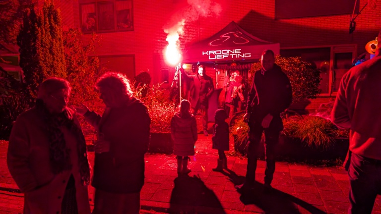 Nachtelijke scène met mensen voor een verlichte tent, waarbij een persoon een fakkel vasthoudt die een rood licht verspreidt.