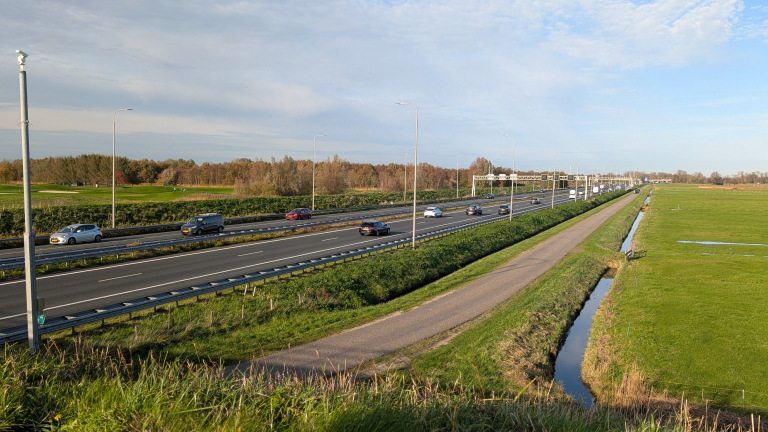 Drukke snelweg met auto's onder een hemel met lichte wolken, omgeven door groene velden en een smalle sloot; lantaarnpalen langs de weg.