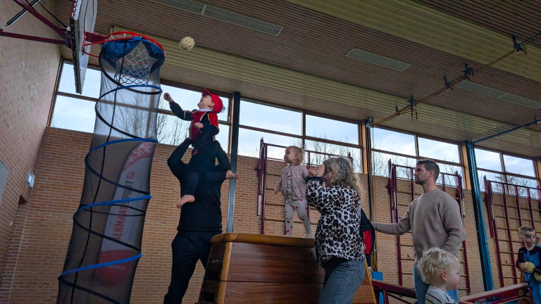 Kinderen en volwassenen in een gymzaal, waarbij een kind op de schouders van een volwassene een bal probeert te gooien in een basket met een lange verenstructuur eronder.