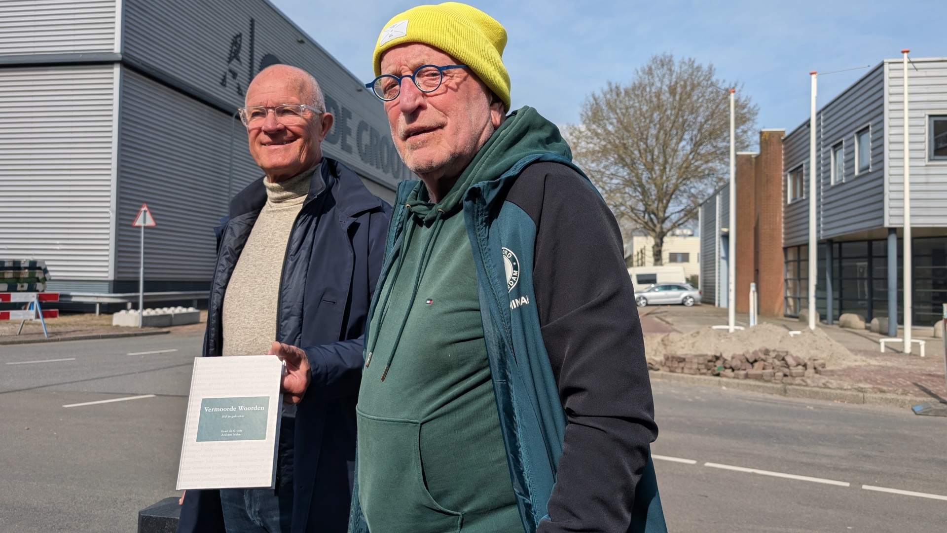 Twee mannen staan buiten voor een kantoor, één draagt een gele muts en houdt een bord met de tekst "Vermoorde Woorden".