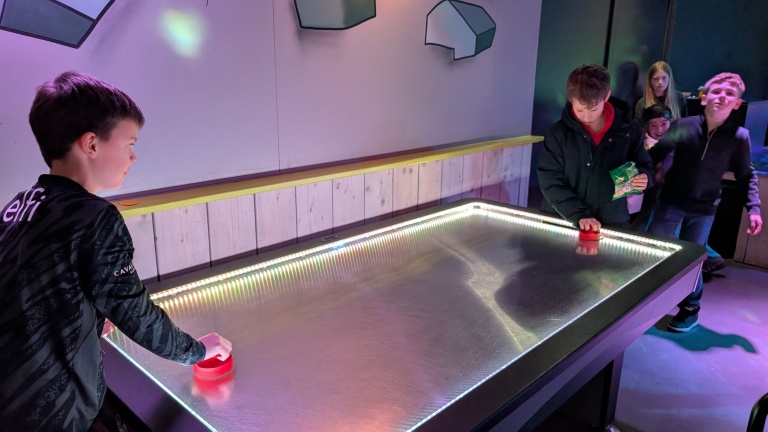 Twee jongens spelen airhockey op een verlicht tafelblad, terwijl anderen toekijken.