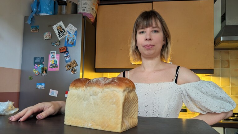 Vrouw in een keuken met een wit bloesje naast een versgebakken brood op het aanrecht, met koelkast en magneten op de achtergrond.