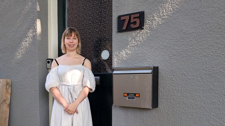 Vrouw in een witte jurk staat voor een grijze muur met een huisnummer 75 en een metalen brievenbus.