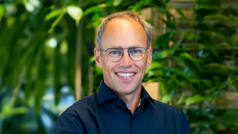 Een man met een bril en een donkerblauw overhemd glimlacht voor een achtergrond met groene planten.