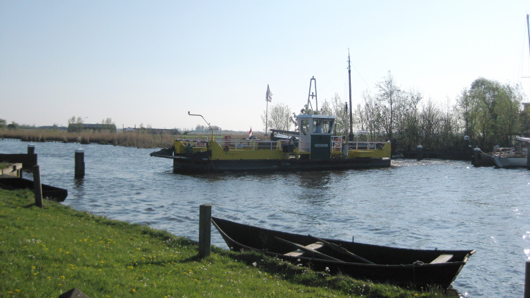 Een veerpont vaart over een rivier, omringd door grasland en bomen onder een heldere lucht.