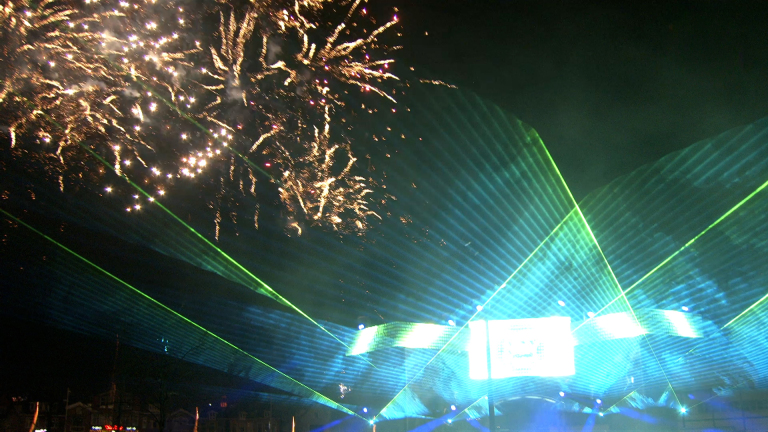 Vuurwerk en groene lasers verlichten de nachtelijke lucht boven een stadsgebied.
