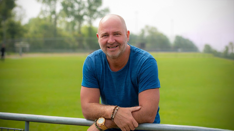 Man in a blauw T-shirt leunt op een hek met een groene grasveld op de achtergrond.