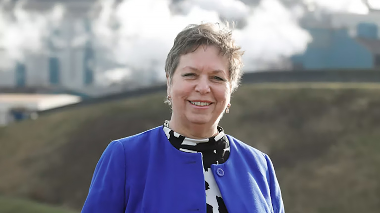 Portret van een glimlachende vrouw in een blauwe blazer, met op de achtergrond een rook uitstotende industrie