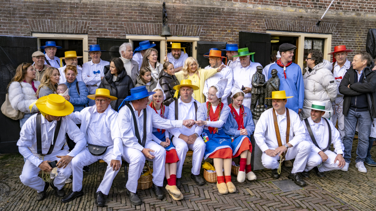 Groep mensen in traditionele Nederlandse kleding, sommigen dragen felgekleurde hoeden buiten een bakstenen gebouw.