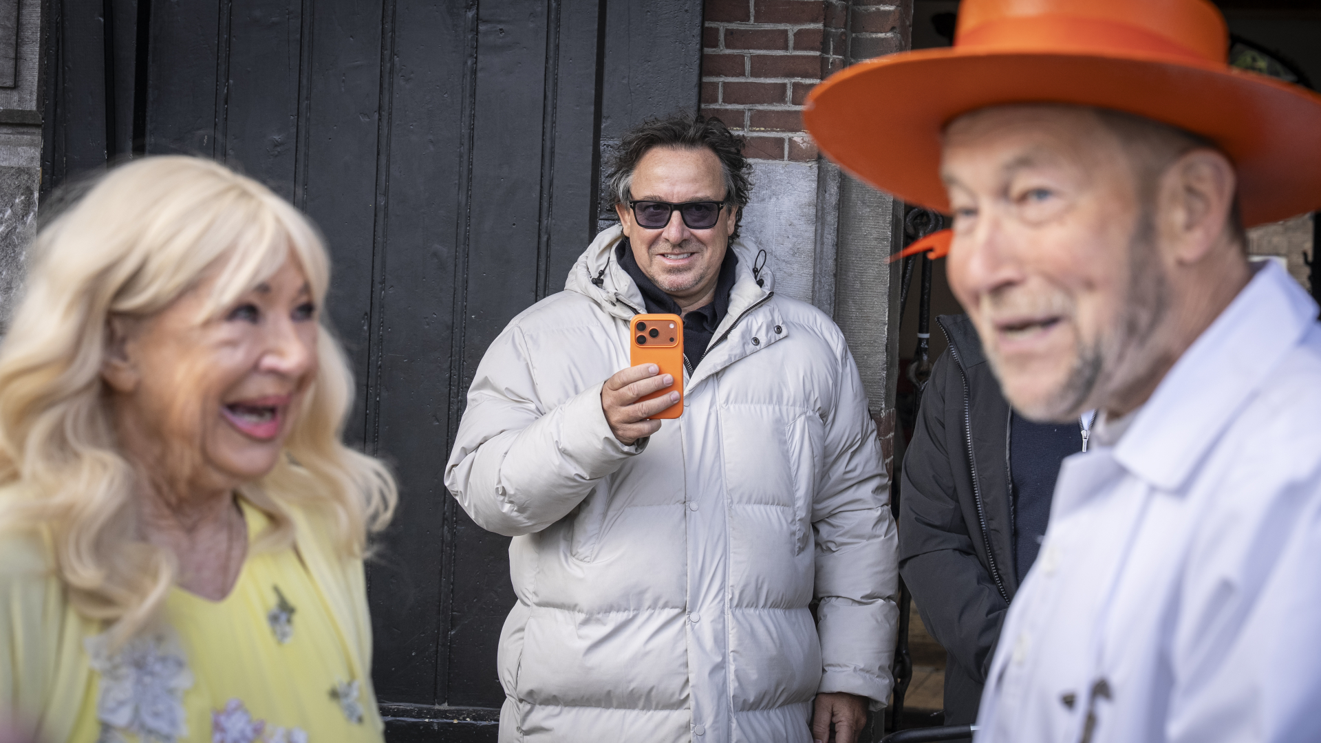 Een man in een witte jas maakt een foto van een lachende vrouw en een man met een oranje hoed.