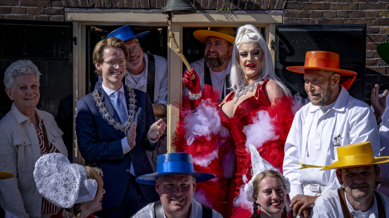 Groepsfoto met mensen in kleurrijke kleding en hoeden, naast een dragqueen in rood met witte veren en een lachende man met een ambtsketen.