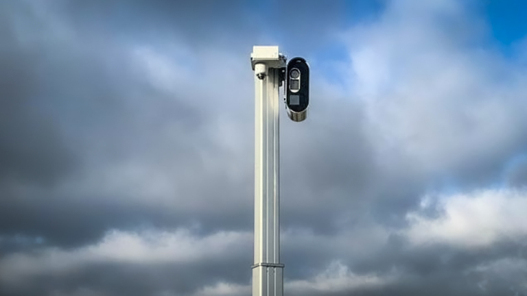 Een hoge metalen paal met een beveiligingscamera erop, tegen een achtergrond van bewolkte lucht.