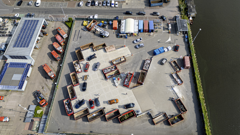 Luchtfoto van een milieupark met verschillende afvalcontainers, auto's en een gebouw met zonnepanelen op het dak.