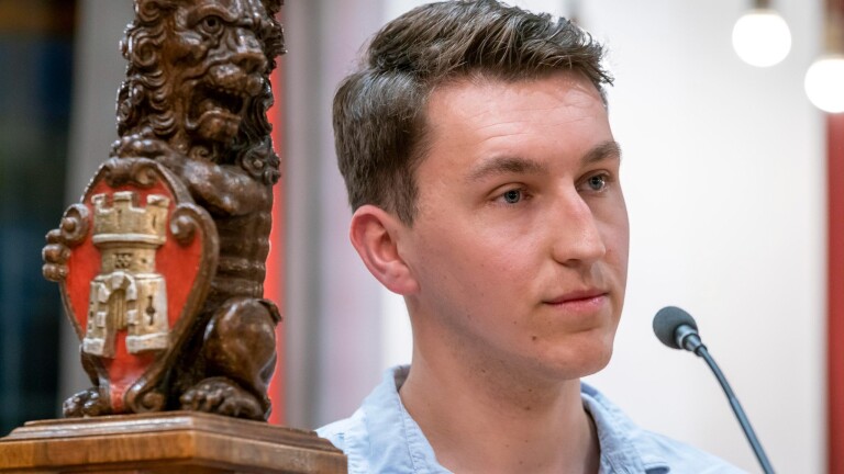 Jonge man bij een microfoon, met naast hem een houten leeuwbeeld met rood schild en kasteelwapen