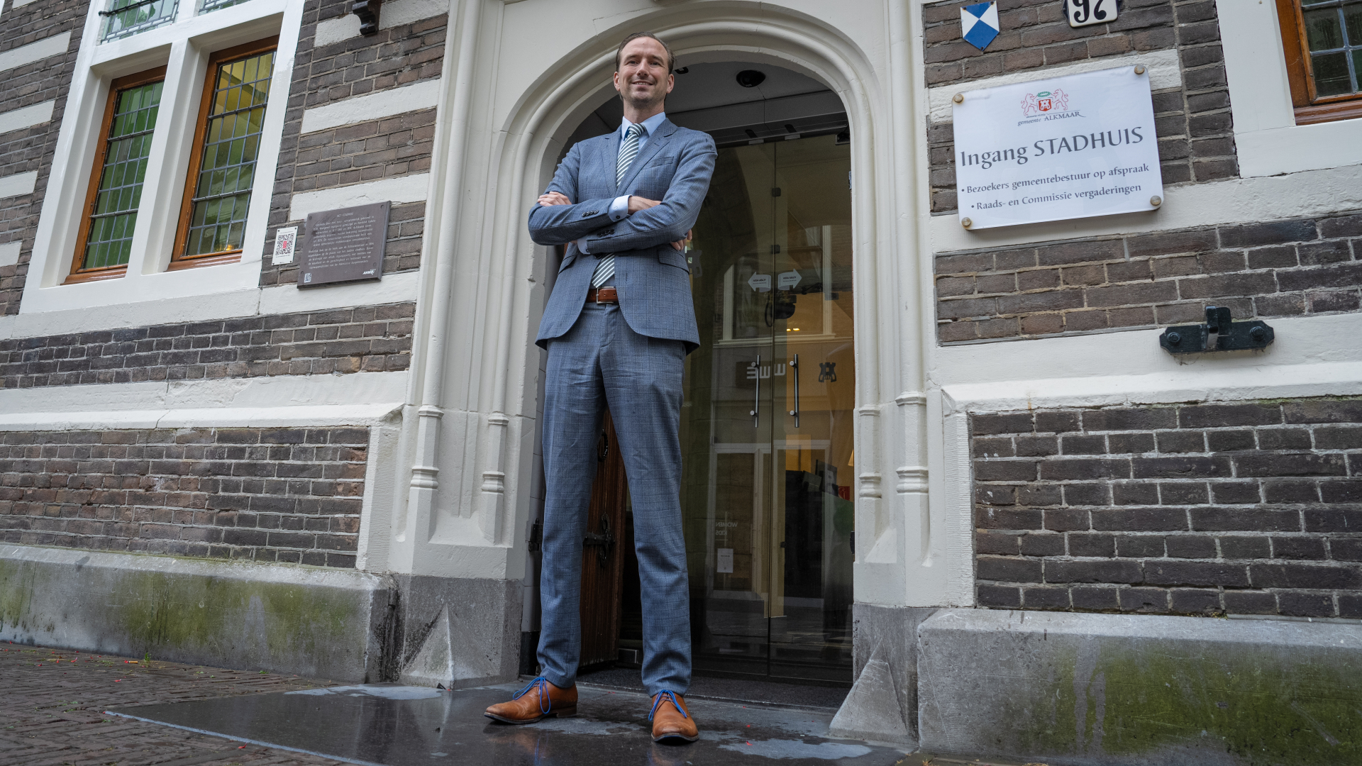 Man in grijs pak staat met gekruiste armen voor de ingang van het stadhuis van Alkmaar.