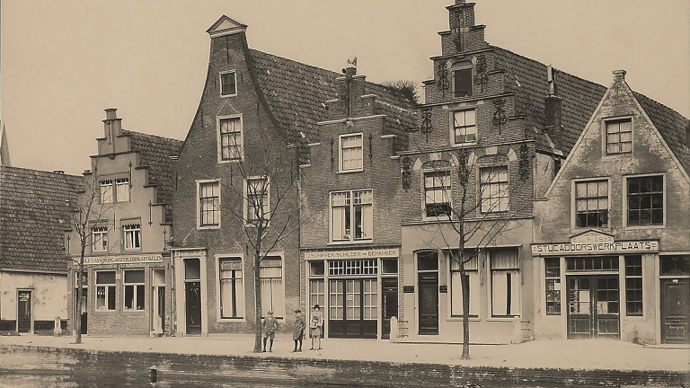 Straatbeeld met oude, traditionele Nederlandse gevels en een aantal mensen die op straat staan.