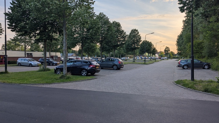 Parkeerplaats in de avondschemering met meerdere geparkeerde auto's en bomen langs de straat aan de linkerkant.