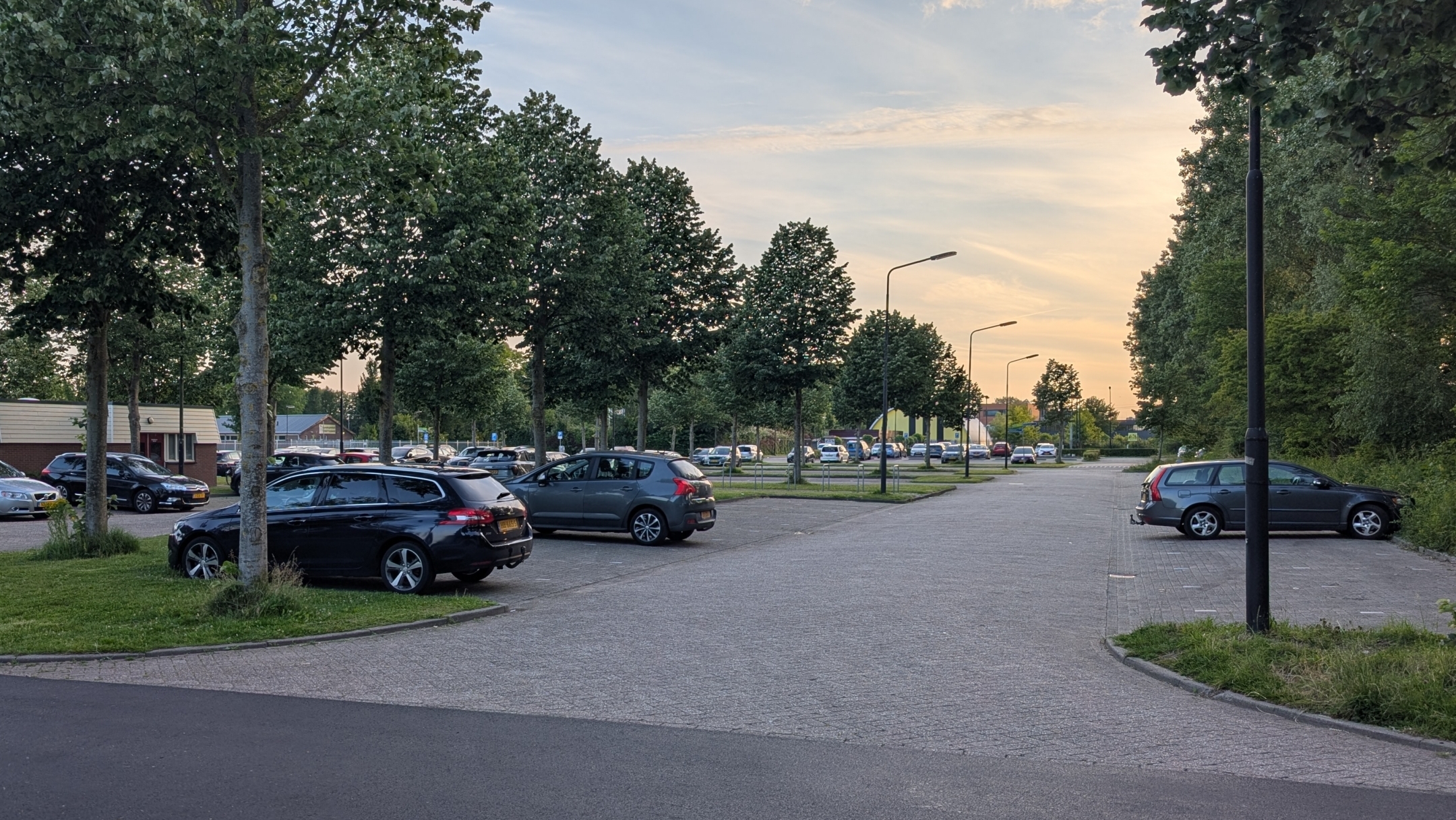Parkeerplaats in de avondschemering met meerdere geparkeerde auto's en bomen langs de straat aan de linkerkant.