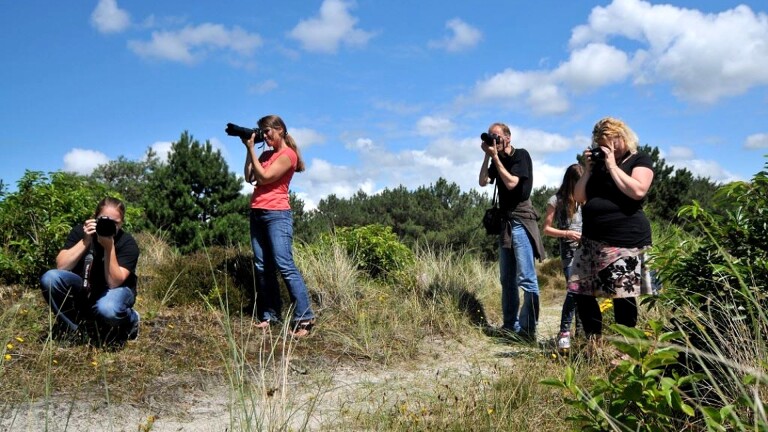 Groep mensen met camera's maakt foto's in een natuurlijke omgeving met gras en bomen, onder een blauwe lucht met wolken.