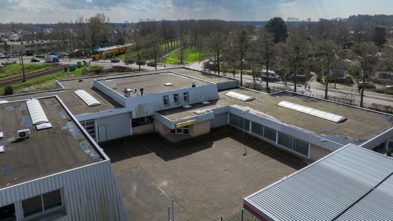 Een groot, utilitair gebouw met een plat dak, gelegen naast een treinspoor met een passerende trein en groene omgeving.