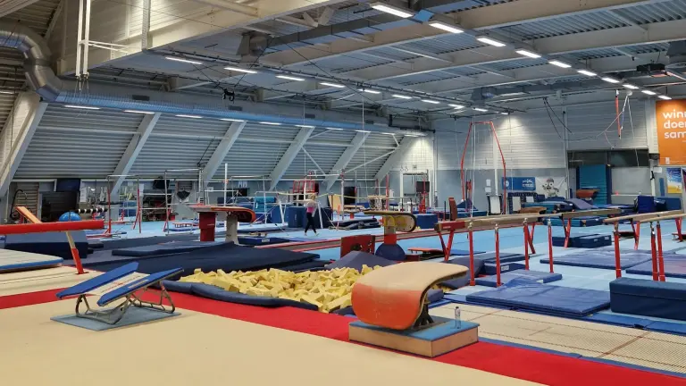 Binnenzicht van een gymnastiekhal met diverse oefeningenapparatuur zoals een springplank, een schuimrubberbak, ringen, balansbalken en matten.