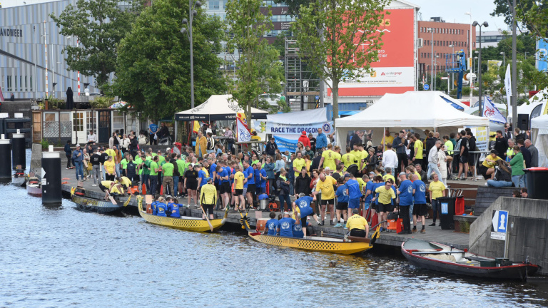 Mensen verzamelen zich bij drakenboten langs een kade, klaar voor een race; kleurrijke shirts en banners zijn zichtbaar.