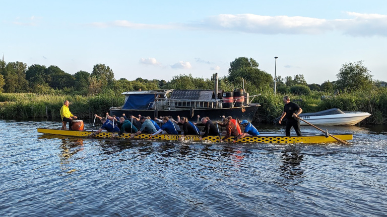 Mensen roeien in een gele drakenboot op een rivier, met groene oevers en boten op de achtergrond.