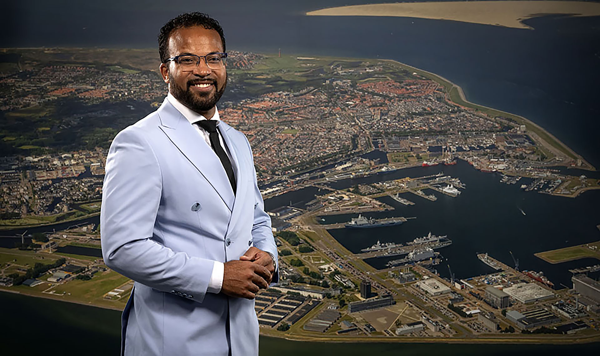 Man in een lichtblauw pak staat voor een luchtfoto van een haven en stad.