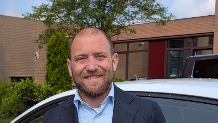 Een lachende man met korte baard en kaal hoofd, gekleed in een donkerblauw pak met een lichtblauw overhemd, staat buiten voor een auto en een gebouw met bakstenen en grote ramen.