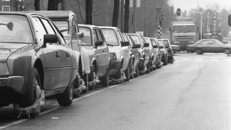 Rij geparkeerde auto’s met wielklemmen langs een straat, met passerend verkeer op de achtergrond