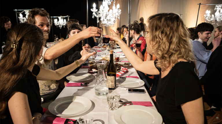Mensen zitten aan een lang gedekte tafel en proosten met glazen wijn, onder elegante kroonluchters.