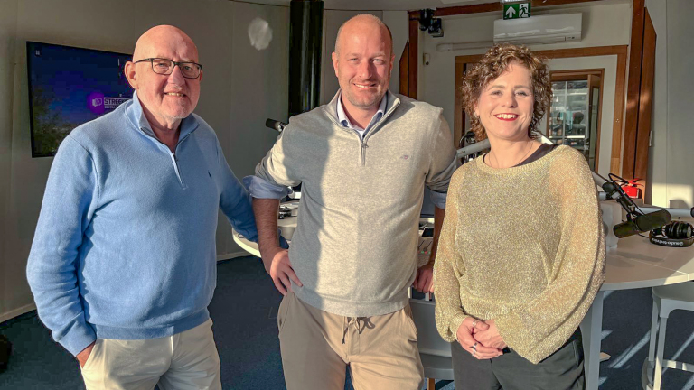 Drie mensen poseren in een radiostudio, met microfoons en mengpaneel op de achtergrond